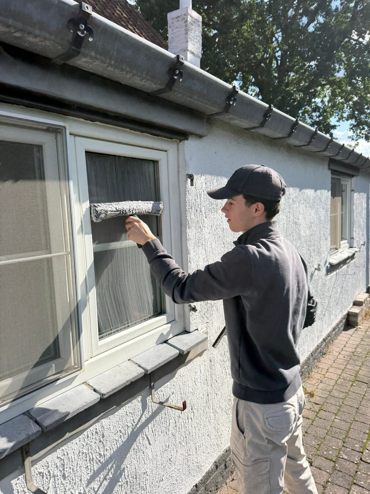 Thibeau haalt een gevelraam schoon met een wasser, gezien vanaf de stoep.